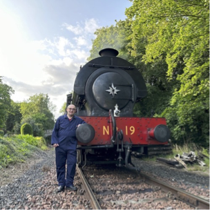 Andrew Sage standing beside the railway track leaning on the front buffer of a steam locomative amongst the trees.